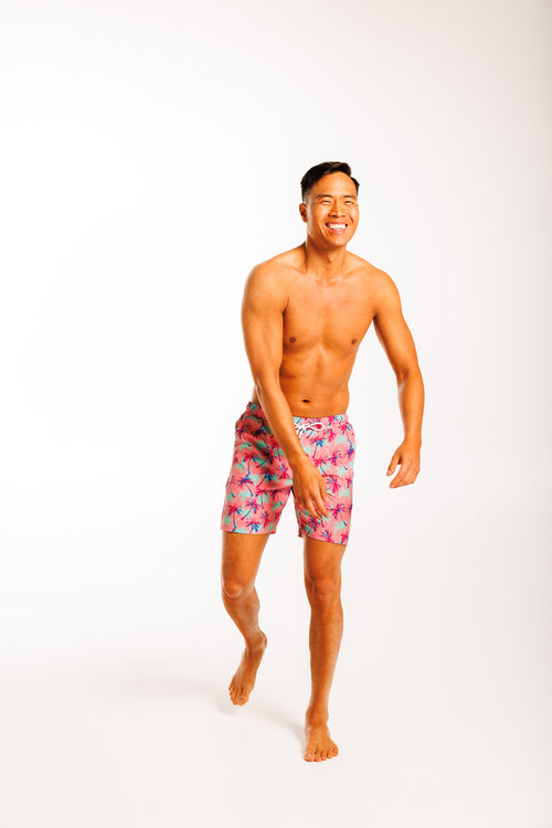 Men's swim shorts featuring a tropical print with falling palm trees against a pink background. The shorts have an elastic waistband with a drawstring and a back zipped pocket, designed for a relaxed, beach-ready look.