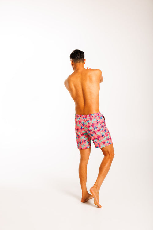 Men's swim shorts featuring a tropical print with falling palm trees against a pink background. The shorts have an elastic waistband with a drawstring and a back zipped pocket, designed for a relaxed, beach-ready look.